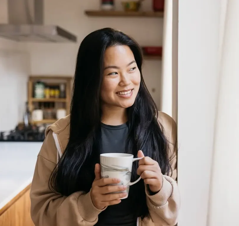 Woman standing indoors with a cup of coffee, smiling warmly in a relaxed comfortable setting