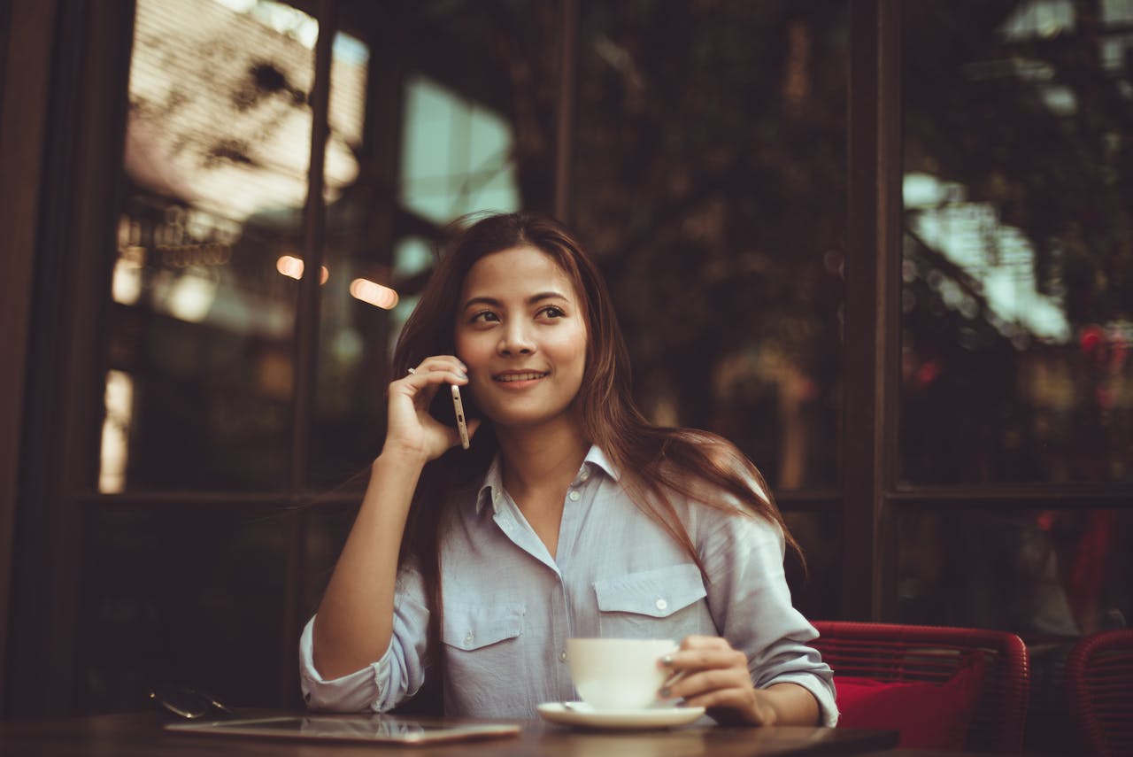 Asian woman talking on the phone at a café while holding a coffee cup.