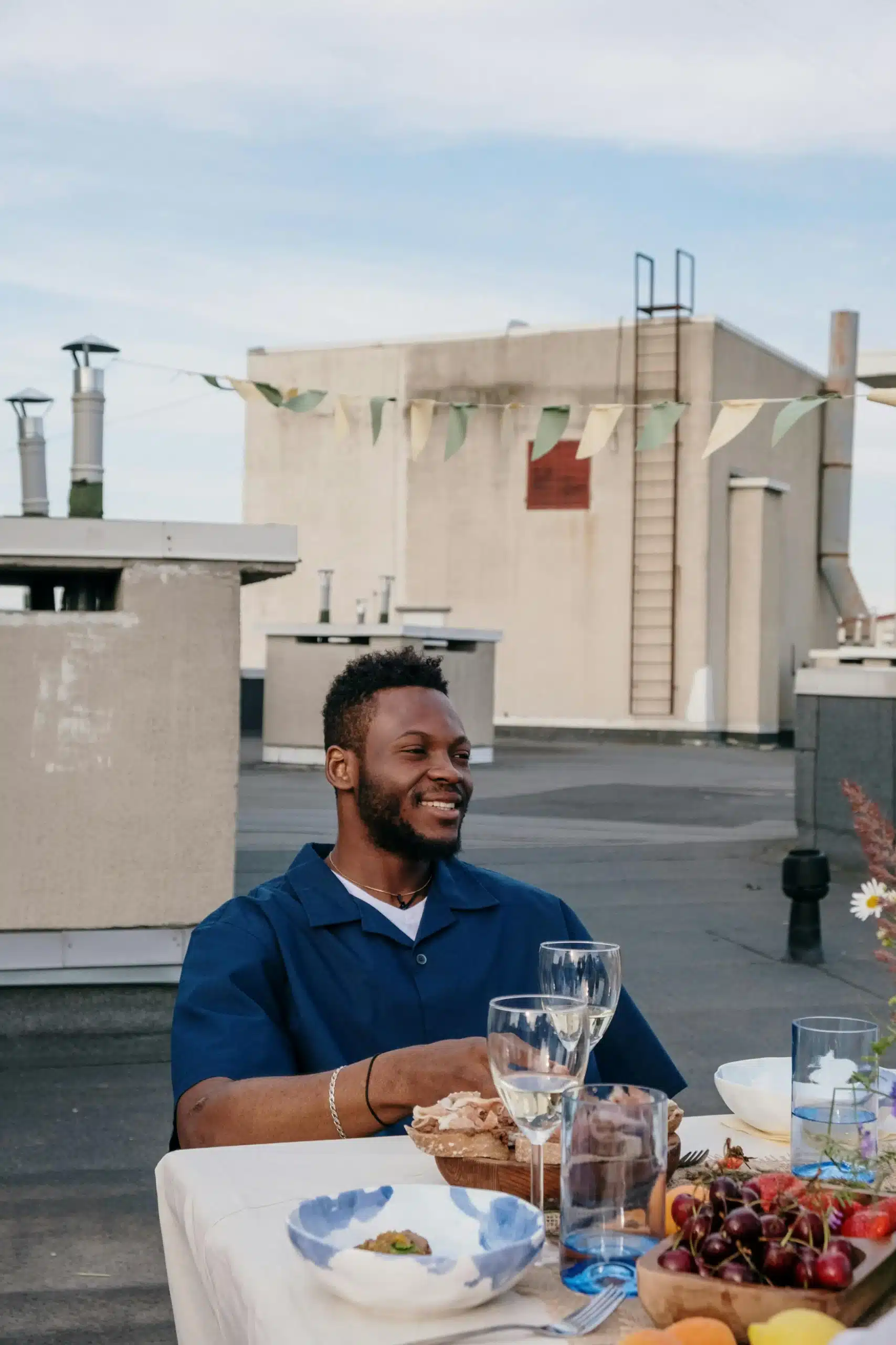 African American man seated on a rooftop with a table, glasses, and food, relaxed posture in a social setting