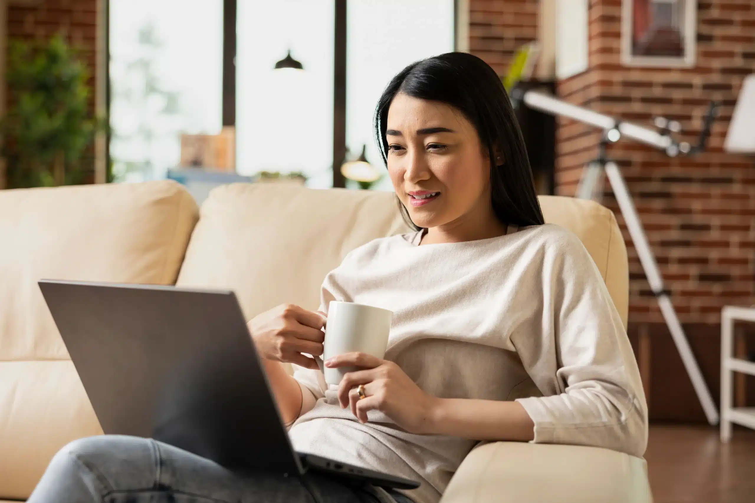 Asian woman sitting with a laptop and coffee, focused on the screen in a calm, comfortable workspace