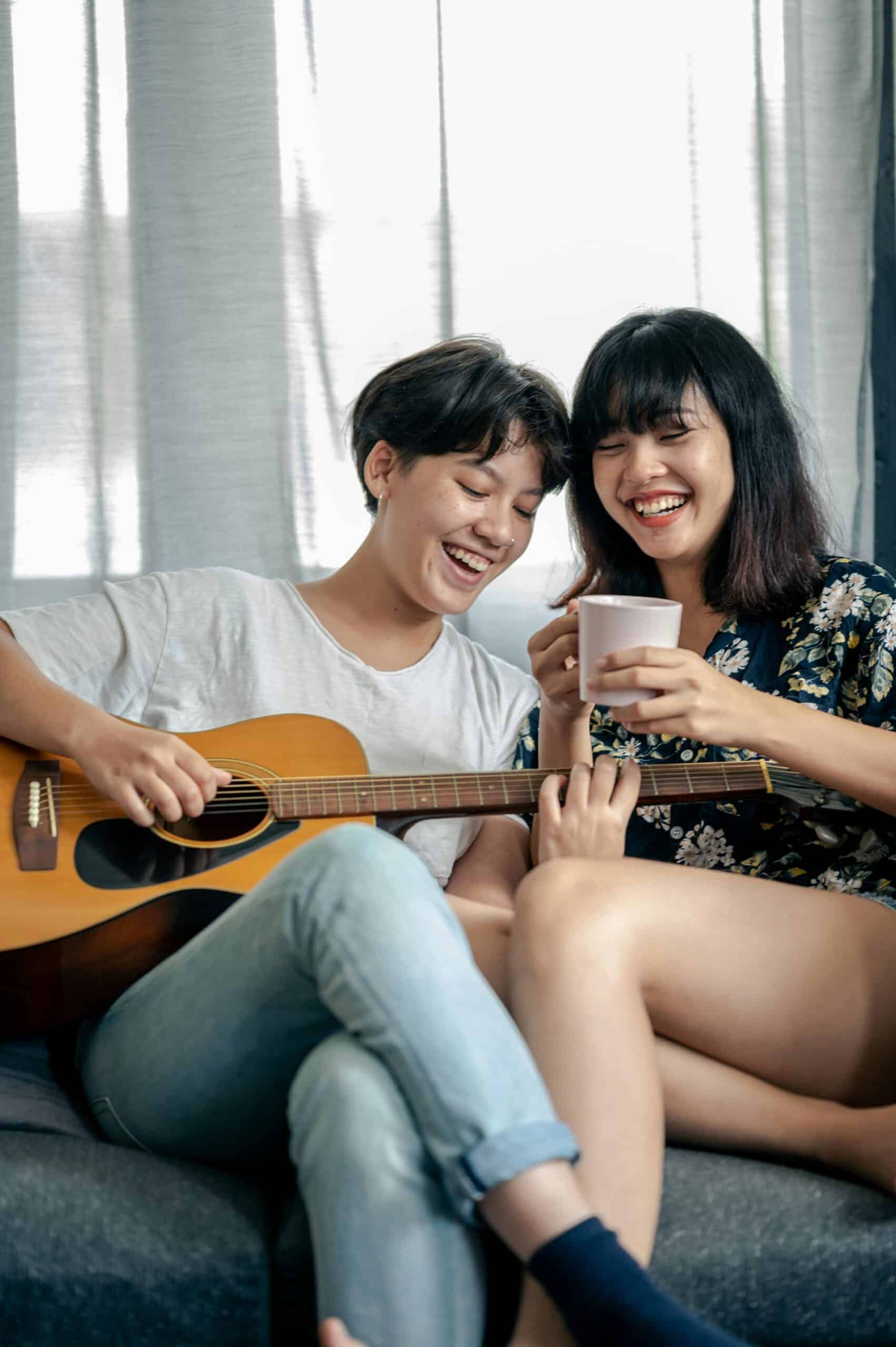 Two girls smiling, one holding coffee and the other playing guitar, symbolizing connection, joy, and shared experience.