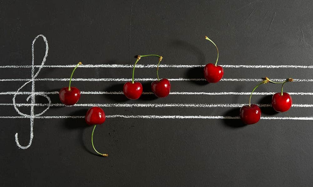 Red cherries arranged as musical notes on white chalk staff lines with a treble clef on a black background.