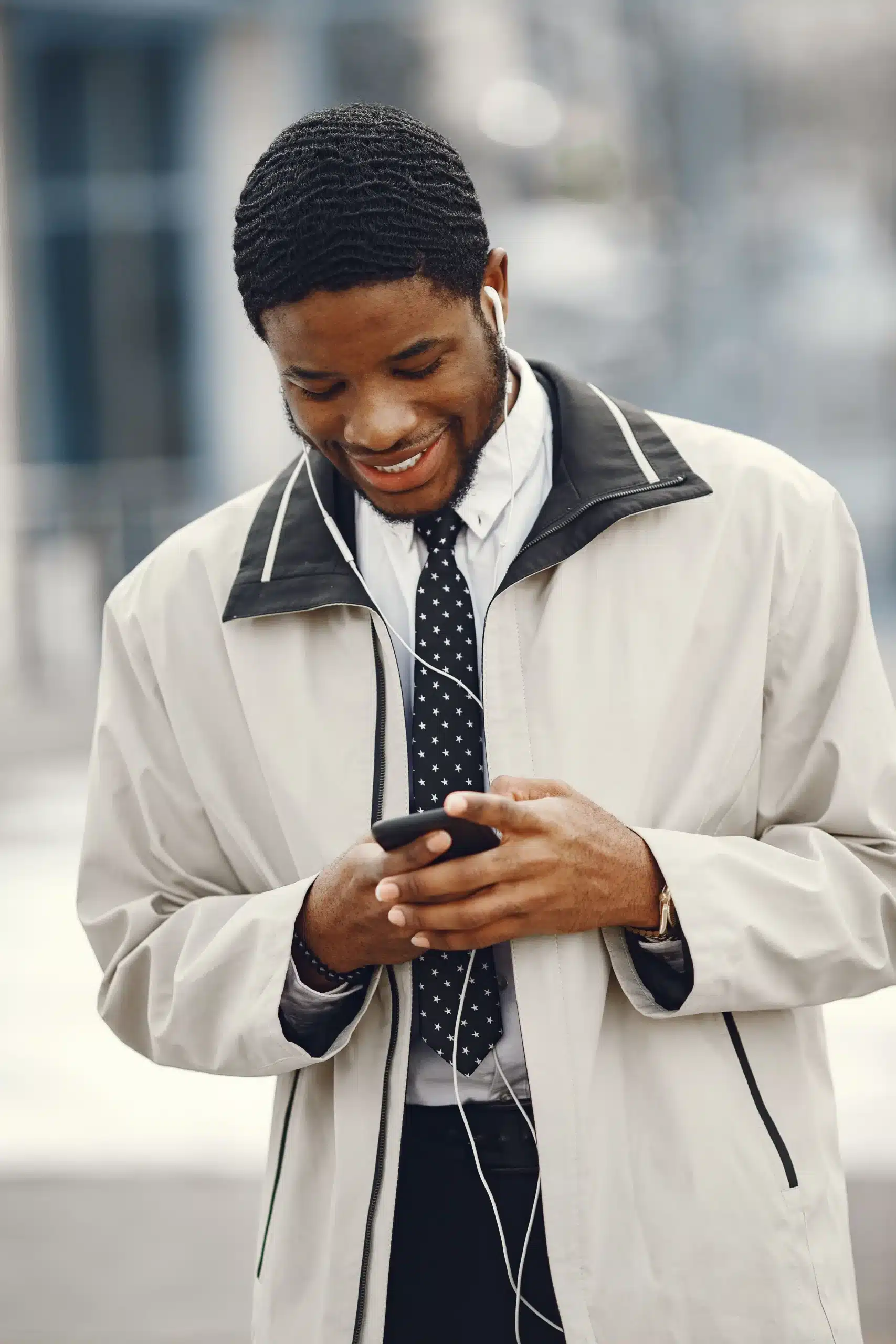 Smiling young man wearing a light beige jacket and tie, typing on his smartphone with wired earphones in.