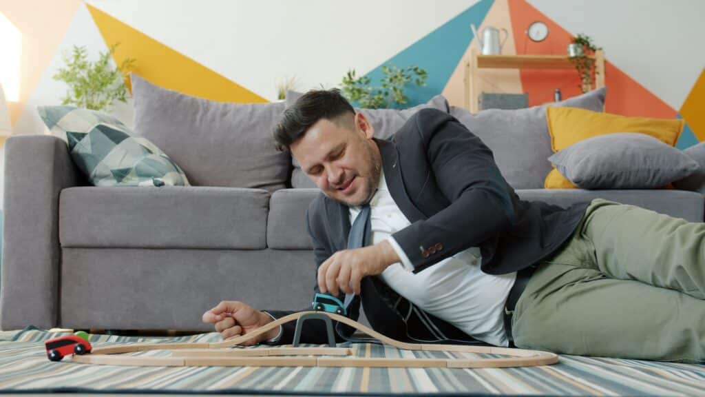 A father in a suit and tie laying on the living room floor playing with a wooden toy train set.