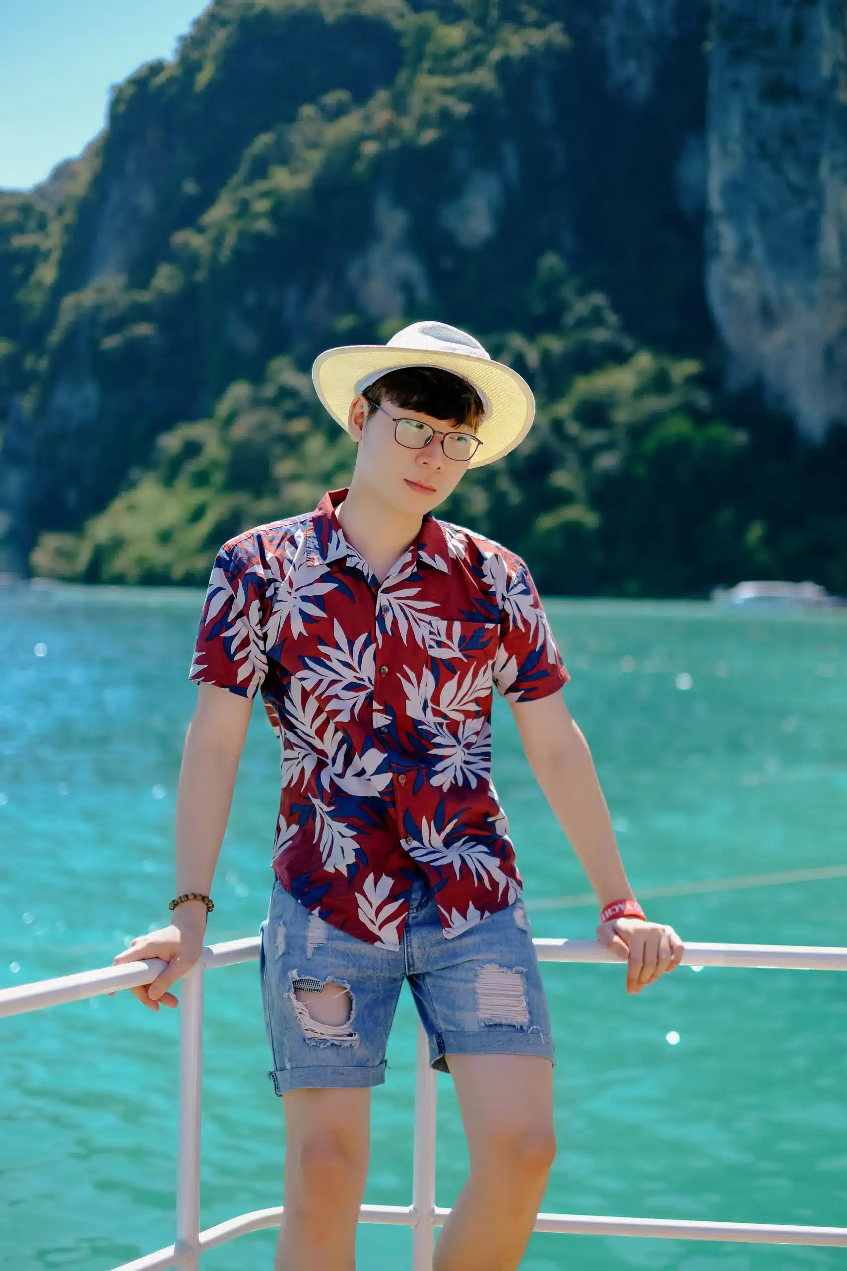 A young man wearing glasses, a sun hat, a red floral tropical shirt, and denim shorts standing on a boat railing with clear turquoise water and lush green hills in the background.