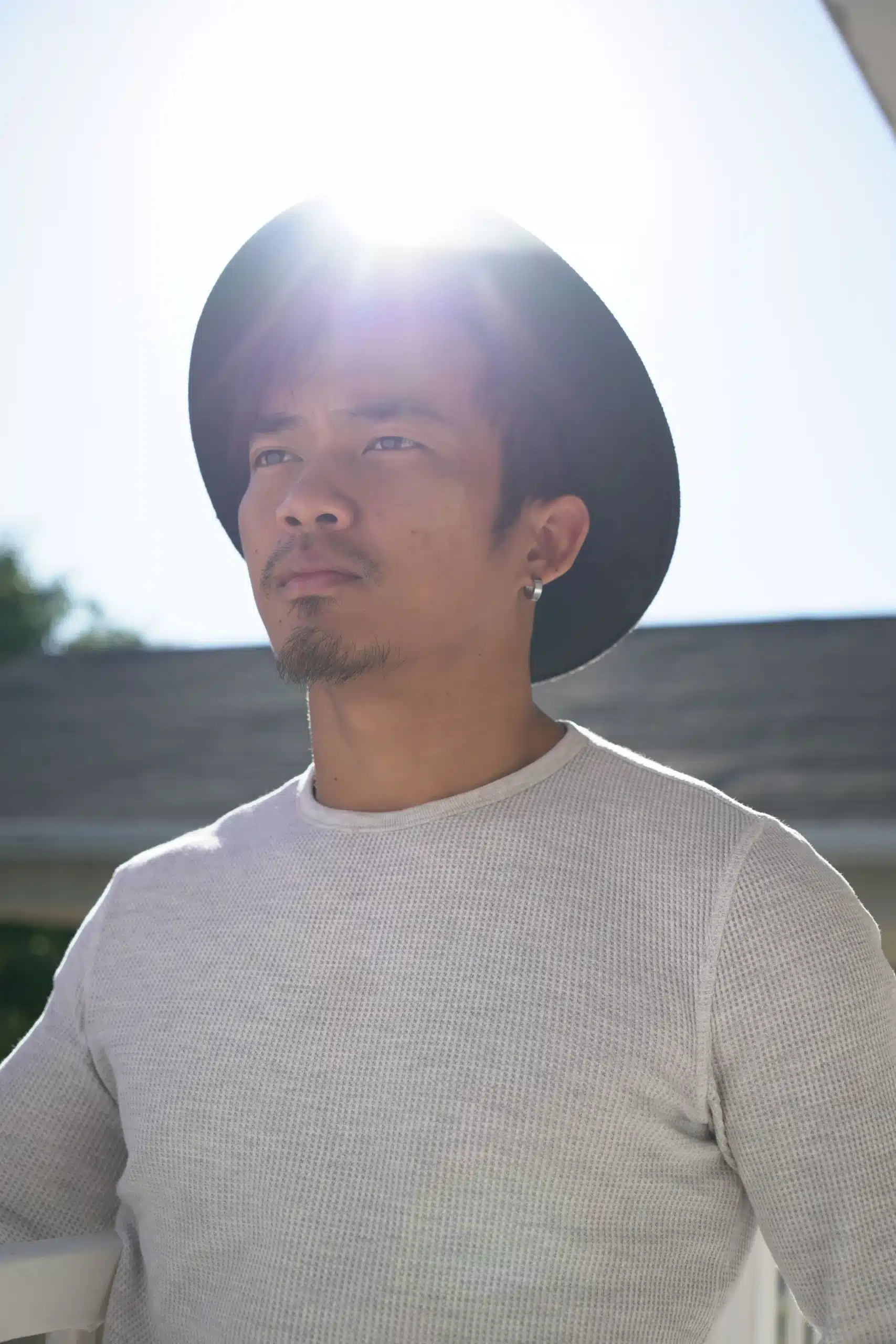 A portrait of a man with facial hair wearing a dark wide-brim hat and a off-white waffle-knit long-sleeve shirt, standing outdoors with bright sunlight behind him.