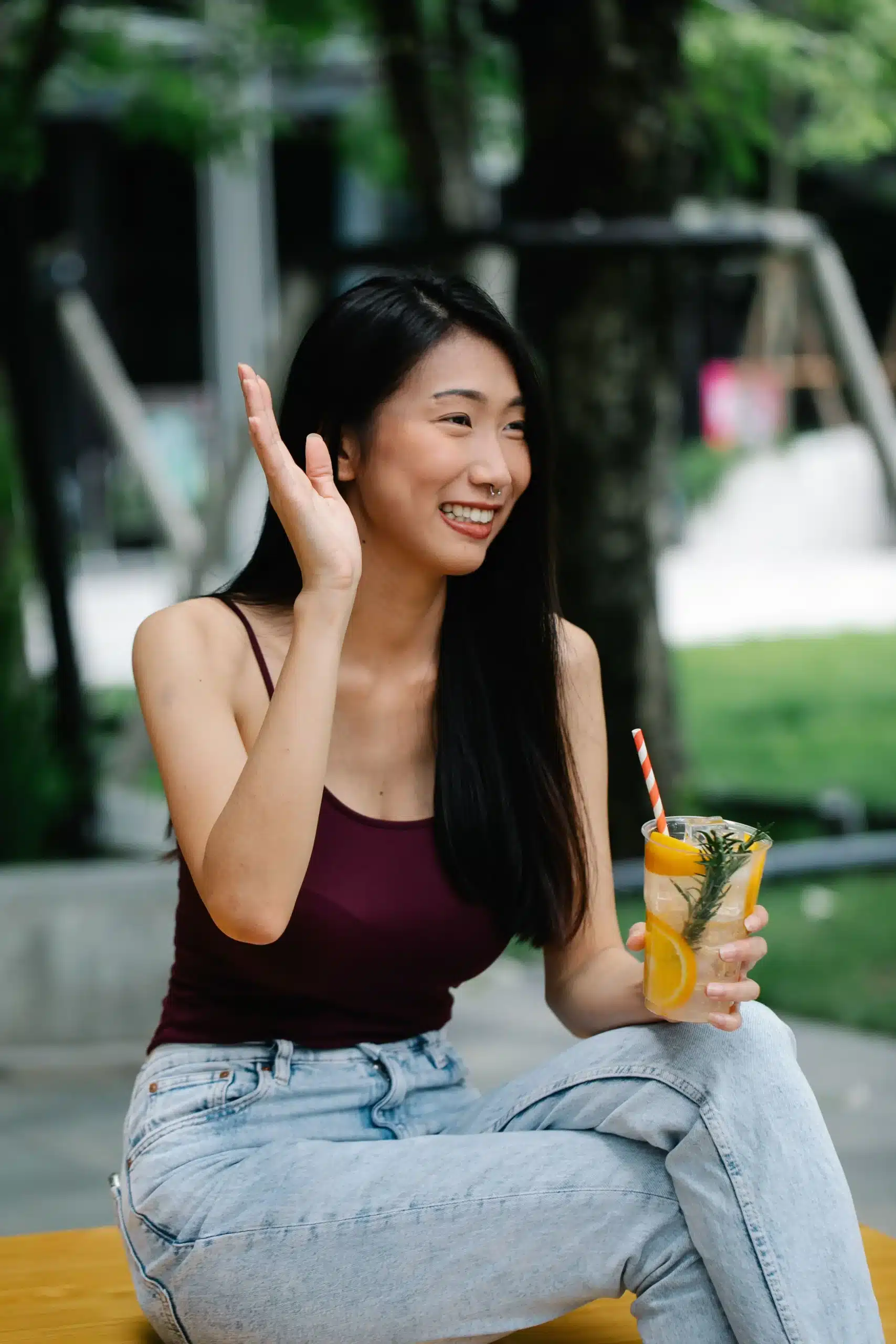 A smiling young woman with dark hair wearing a maroon tank top and jeans sitting outdoors, waving her hand while holding a clear plastic cup filled with an iced citrus drink garnishing orange slices and rosemary.