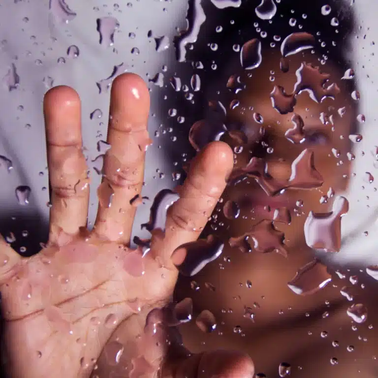 Hand pressed against a wet glass surface covered in water droplets with a blurred face visible in the background.