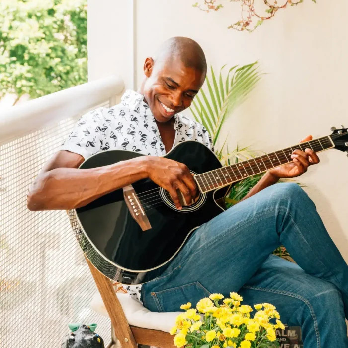 African man seated outdoors playing guitar in daylight, smiling as he connects with music in a relaxed setting