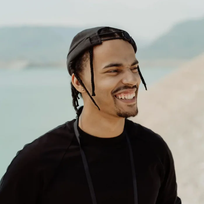 Close-up of a smiling man with braids and a black cap, outdoors